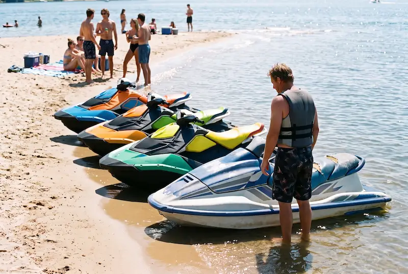 Rider beside a faded older jet ski at a sandbar, glancing at a row of glossy newer skis